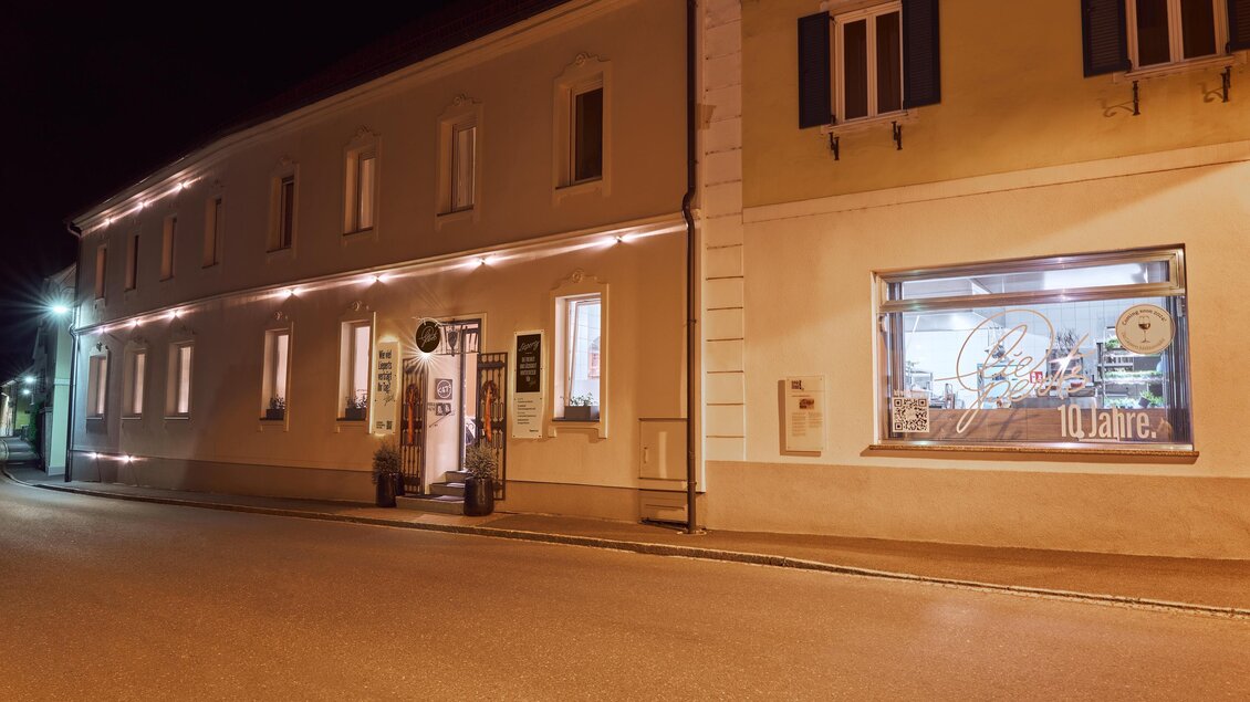 Eine ruhige Straße bei Nacht mit einem beleuchteten Gebäude. Das Fenster zeigt eine ansprechende Auslage.