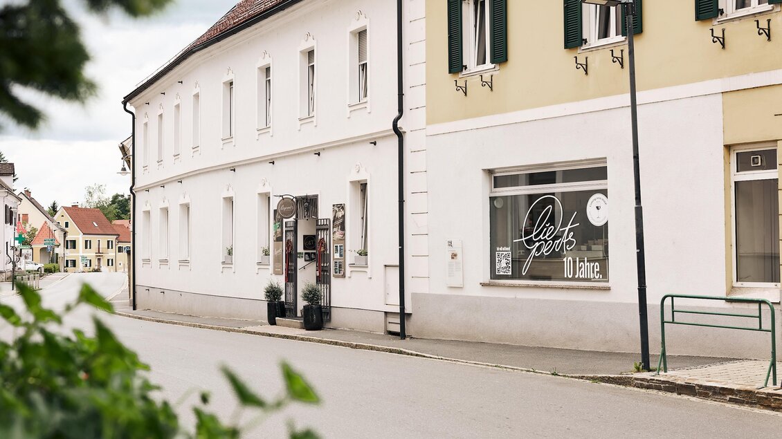 Eine ruhige Straßenszene mit Gebäuden in hellen Farben. Im Vordergrund ist ein Geschäft mit einer großen Fensterfront zu sehen.