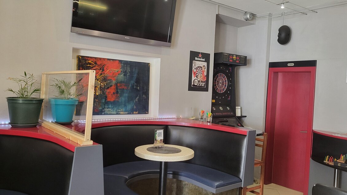 Ein gemütliches Café mit Sitzgelegenheiten und einem kleinen Tisch. Im Hintergrund sind ein Fernseher und eine Dartscheibe zu sehen. | © TV Hochsteiermark