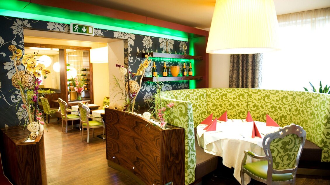 Ein moderner Restaurantbereich mit grüner Polsterung und stilvollen Dekorationen. Auf den Tischen stehen rote Servietten und es herrscht eine angenehme Atmosphäre. | © Lindenwirt - RP photography +