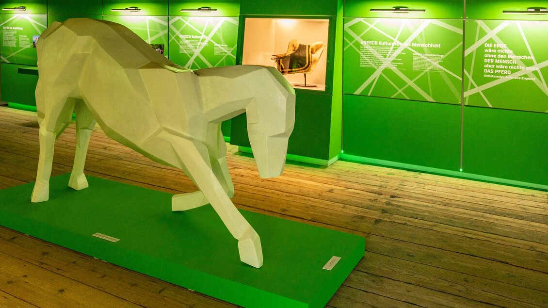Eine moderne, geometrische Skulptur eines Pferdes auf einem grünen Sockel. Im Hintergrund sind farbige Wände und beleuchtete Objekte in einer Ausstellungsumgebung zu sehen. | © SHS-LipizzanergestütPiberGöR - Renè van Bakel