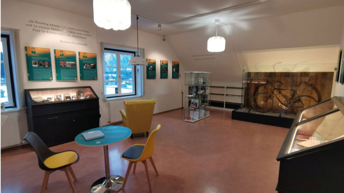 Ein modernes Ausstellungszimmer mit informativen Tafeln an den Wänden. In der Mitte stehen ein runder Tisch mit Stühlen und ein Vitrinenschrank, in dem ein Fahrrad präsentiert wird. | © Kulturfux