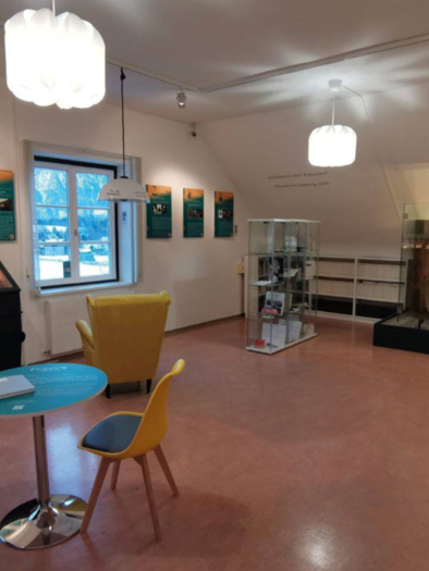 A modern exhibition room with informative panels on the walls. In the center stands a round table with chairs and a display cabinet in which a bicycle is presented. | © Kulturfux