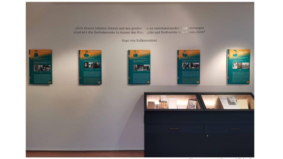 Eine Ausstellung mit Infotafeln an der Wand und einem Vitrinenschrank. Die Wände sind hell gestrichen und die Tafeln sind grün mit Text und Bildern. | © Kulturfux