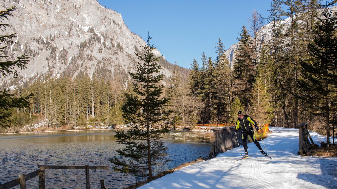Ein Skilangläufer auf einer verschneiten Strecke neben einem See. Im Hintergrund sind hohe Berge und große Bäume zu sehen. | © Eva Rossböck