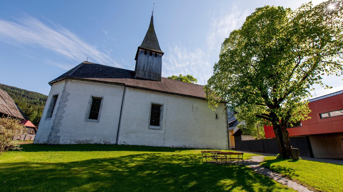 Eine alte Kirche mit einem spitzen Turm steht auf einer grünen Wiese. Im Hintergrund sind Bäume und ein modernes Gebäude zu sehen. | © Holzwelt Murau