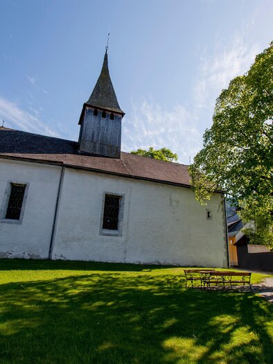 An old church with a pointed tower stands in a green meadow. In the background, there are trees and a modern building. | © Holzwelt Murau