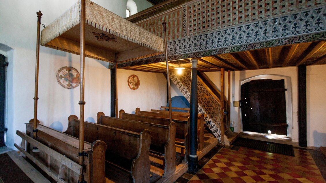 Ein historischer Innenraum mit Holzbänken und einem kunstvoll verzierten Obergeschoss. Der Boden ist mit bunten Fliesen gestaltet. | © Holzwelt Murau