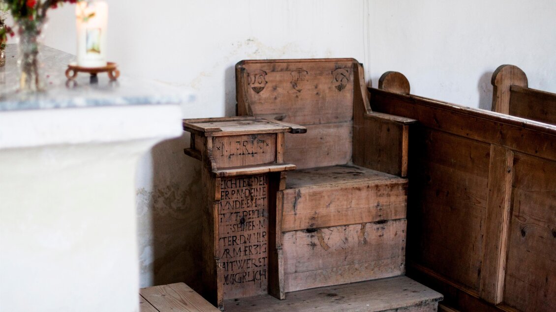 Ein alter Holzstuhl in einer Kirche, mit sichtbaren Kratzern und Inschriften. Der Stuhl steht neben einer hölzernen Kirchenbank. | © Holzwelt Murau