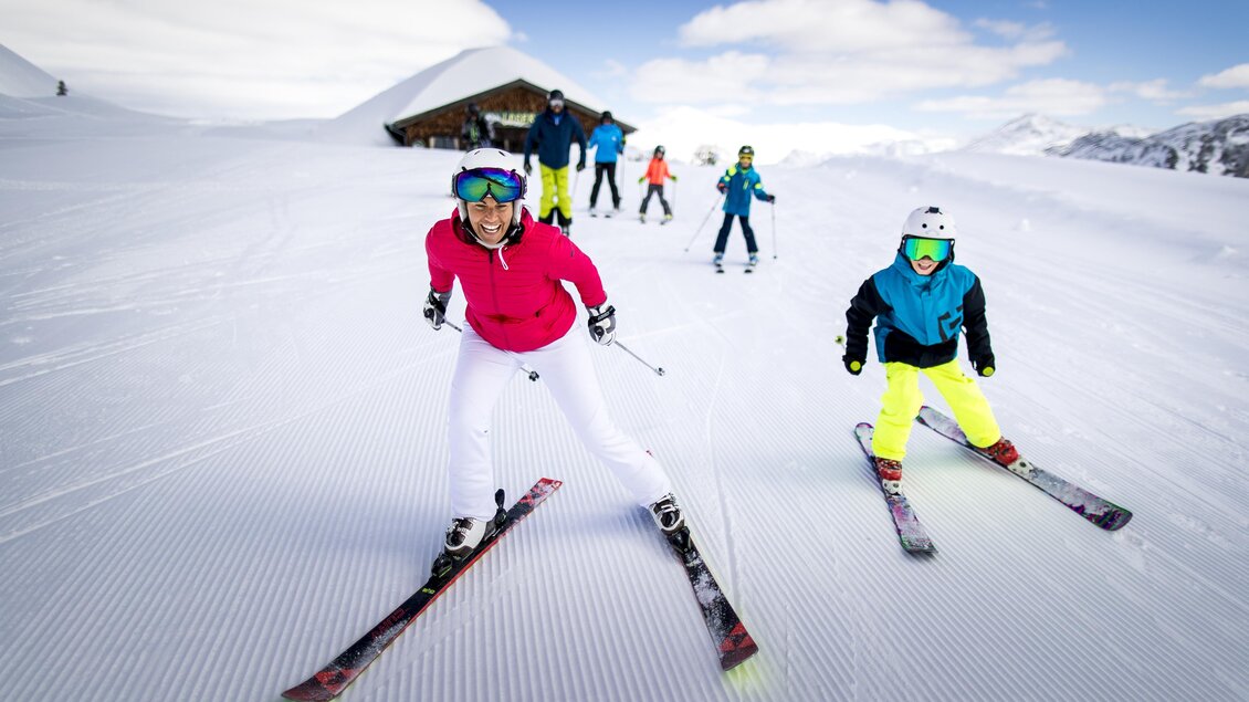 Eine Gruppe von Skifahrern fährt über eine schneebedeckte Piste. Im Hintergrund ist ein Alpenszenario mit einem Skihütte zu sehen. | © TVB Ausseerland Salzkammergut / Tom Lamm