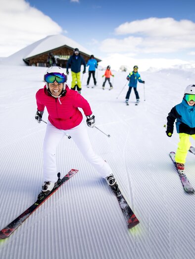Eine Gruppe von Skifahrern fährt über eine schneebedeckte Piste. Im Hintergrund ist ein Alpenszenario mit einem Skihütte zu sehen. | © TVB Ausseerland Salzkammergut / Tom Lamm