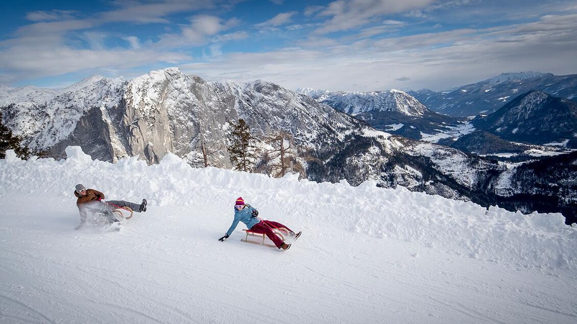 Zwei Personen rodeln auf einer schneebedeckten Piste in den Bergen. Im Hintergrund sind beeindruckende, schneebedeckte Gipfel und ein klarer Himmel zu sehen. | © TVB Ausseerland-Salzkammergut / Tom Lamm