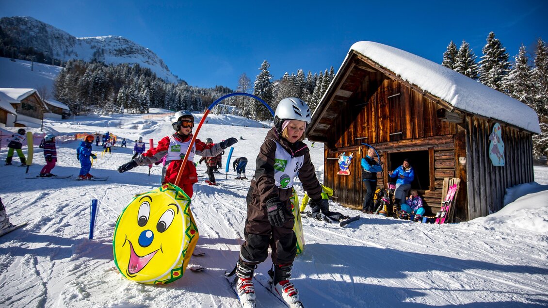 Ein schöner Skikurs mit Kindern auf einer schneebedeckten Piste. Im Hintergrund stehen Holzgebäude und Bäume. | © TVB Ausseerland/Tom Lamm