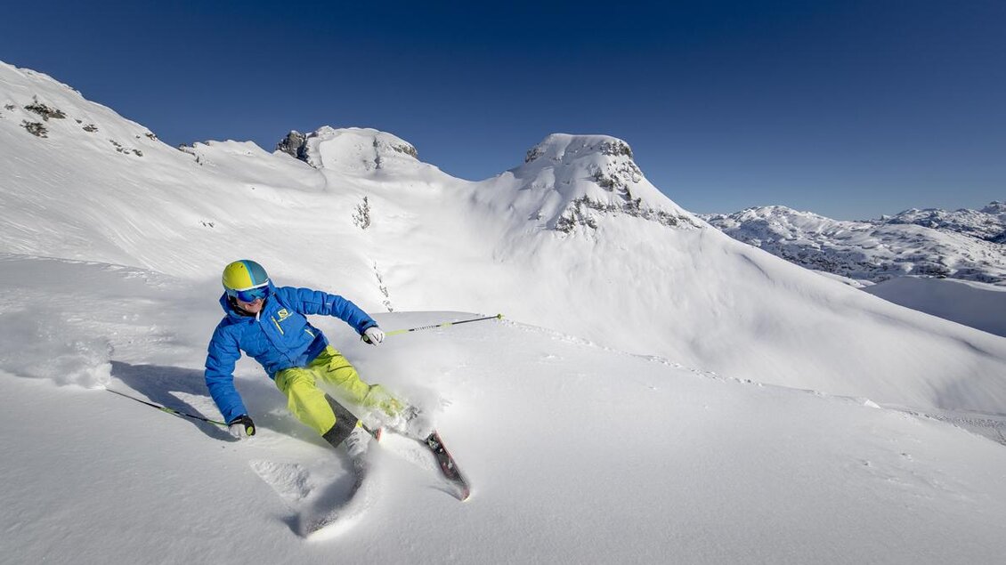 Ein Skifahrer fährt durch frischen, tiefen Schnee. Die Berglandschaft im Hintergrund strahlt unter einem klaren, blauen Himmel. | © TVB Ausseerland-Salzkammergut / Tom Lamm