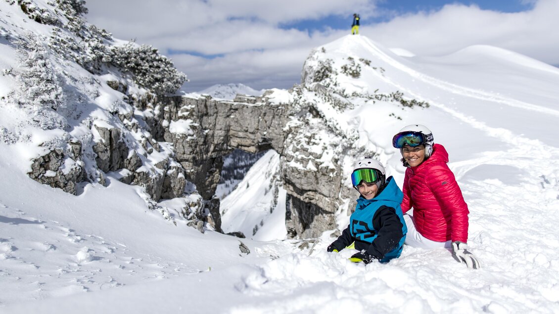 Zwei Kinder spielen im Schnee, während sie bunte Skikleidung tragen. Im Hintergrund ist eine schneebedeckte Landschaft mit Felsen sichtbar. | © Ausseerland Salzkammergut / Tom Lamm
