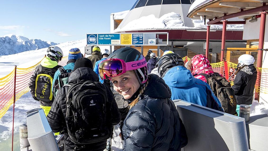 Eine Gruppe Skifahrer steht in einer Schlange vor einer Bergbahn. Im Hintergrund sind schneebedeckte Berge und ein klarer blauer Himmel zu sehen. | © ausseerland-salzkammergut_tom lamm