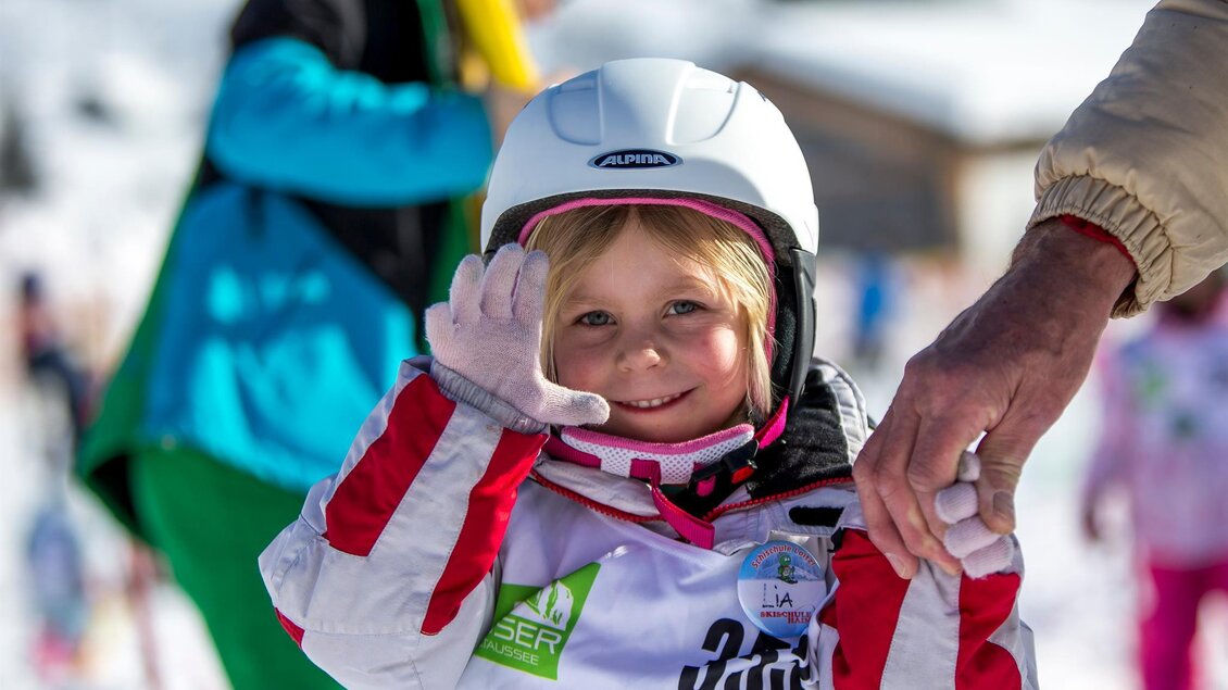 Ein lächelndes kleines Mädchen mit einem Helm und Skikleidung steht im Schnee. Sie winkt fröhlich in die Kamera. | © TVB Ausseerland - Salzkammergut/T. Lamm