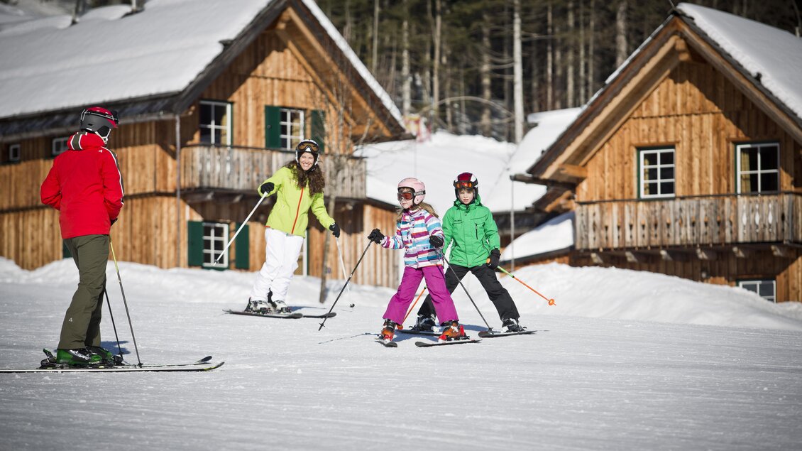 Eine Gruppe von Skifahrern genießt einen sonnigen Tag im Schnee. Im Hintergrund sind gemütliche Holzhütten und ein Wald sichtbar. | © Loser Bergbahnen/mirjageh.com
