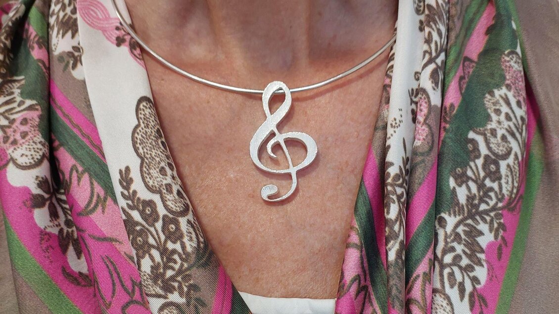 Eine Nahaufnahme eines Halsbands mit einem silbernen Notenschlüssel-Anhänger. Die Person trägt einen bunten Schal mit floralen Mustern. | © Lisa Wagenhofer
