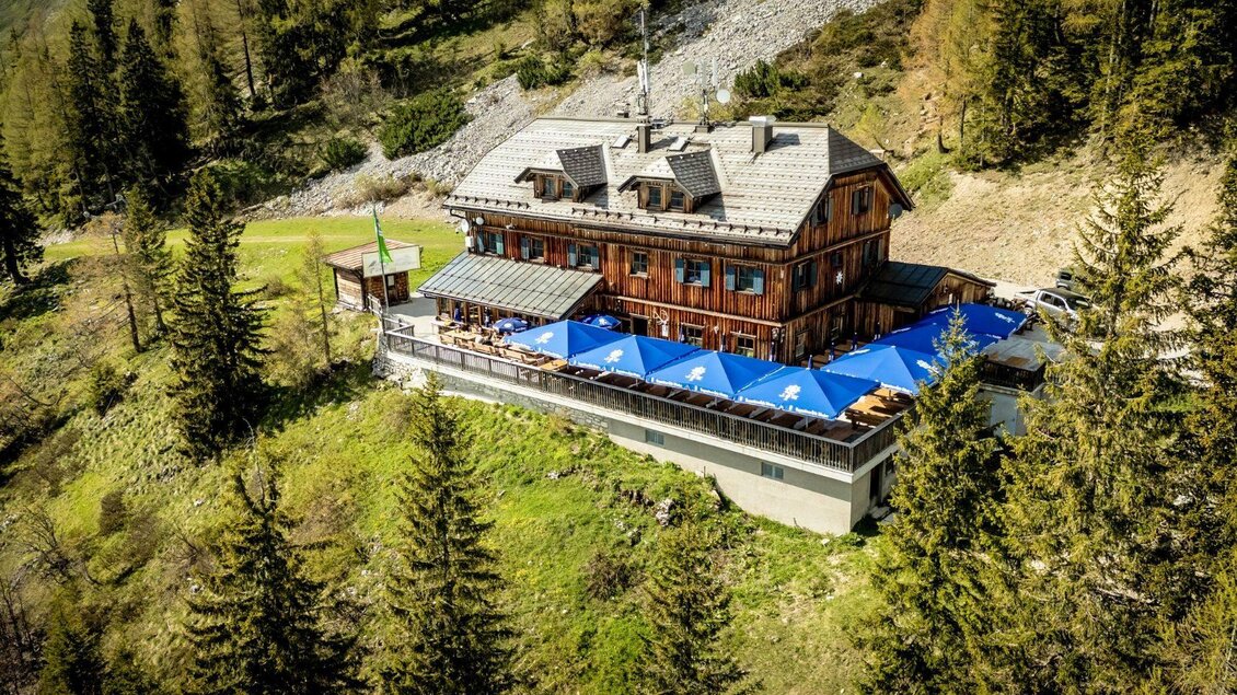 Ein rustikales Chalet umgeben von Bäumen in den Bergen. Die Terrasse mit blauen Sonnenschirmen lädt zum Entspannen ein. | © Chris Gütl