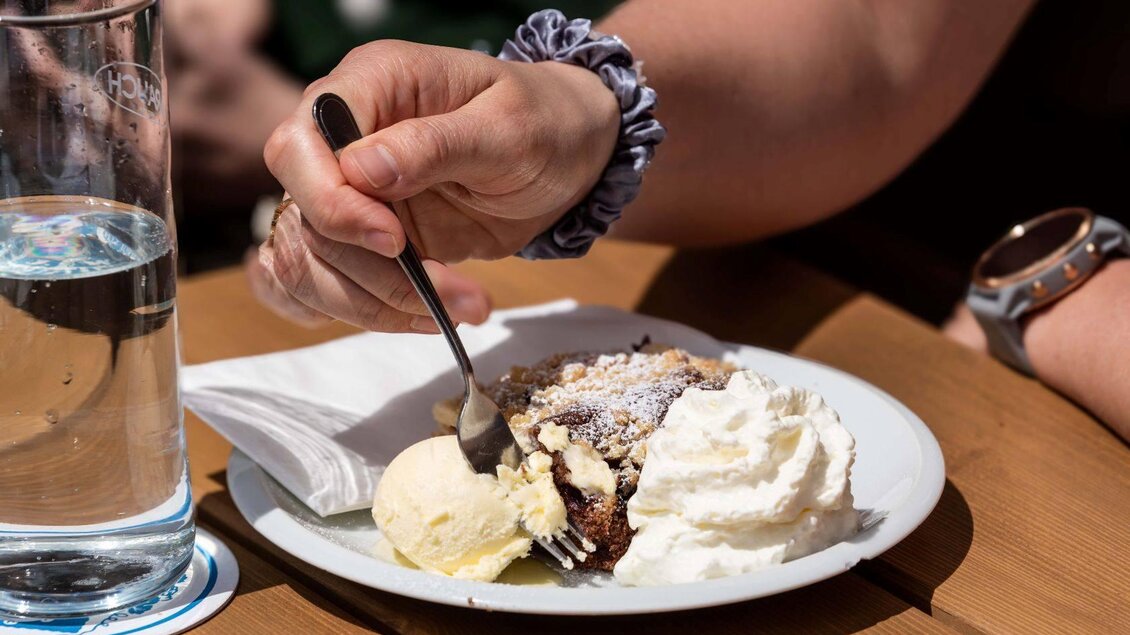 Ein Stück Kuchen mit Sahne und Eis auf einem Teller. Eine Hand hält eine Gabel und bereitet sich darauf vor, den Kuchen zu essen. | ©  Chris Gütl