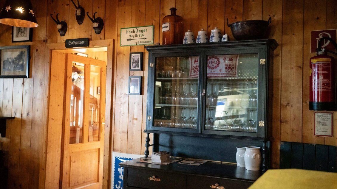 Ein rustikales Zimmer mit Holzvertäfelung, antikem Geschirr und einer Glaskommode. Im Hintergrund ist eine Holztür sichtbar, die zu einem anderen Bereich führt. | © Chris Gütl