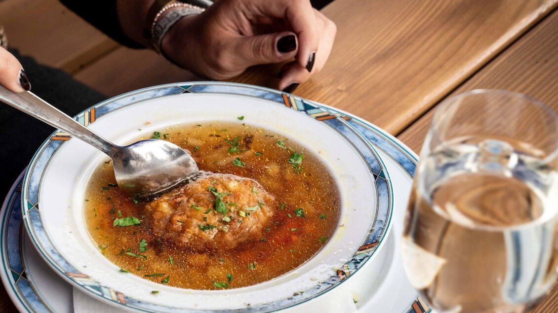Eine Schale mit klarer Suppe und einem Fleischklößchen, garniert mit frischen Kräutern. Neben dem Teller steht ein Glas Wasser. | ©  Chris Gütl