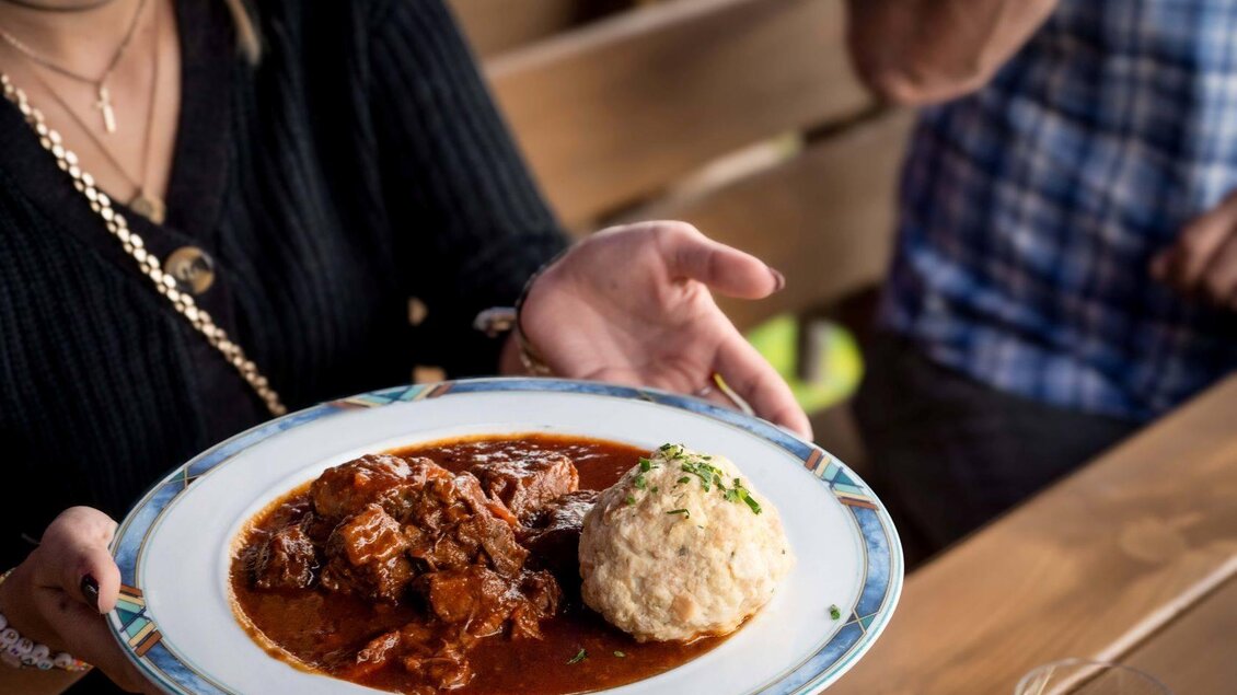 Ein Teller mit herzhaftem Rindfleisch in einer dunklen Sauce und einem Knödel. Der Hintergrund zeigt eine rustikale Tischatmosphäre. | ©  Chris Gütl