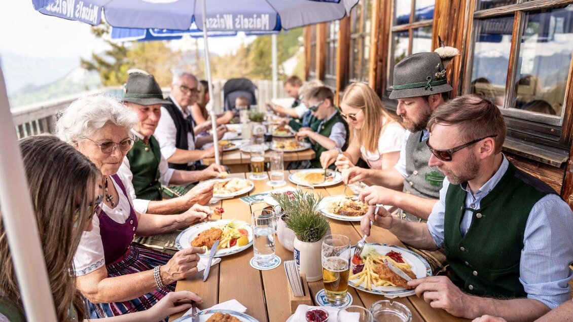 Eine Gruppe von Menschen in traditioneller Kleidung sitzt an einem Tisch im Freien und genießt ein Essen. Es sind Bier und typisch bayerische Gerichte auf dem Tisch angerichtet. | ©  Chris Gütl