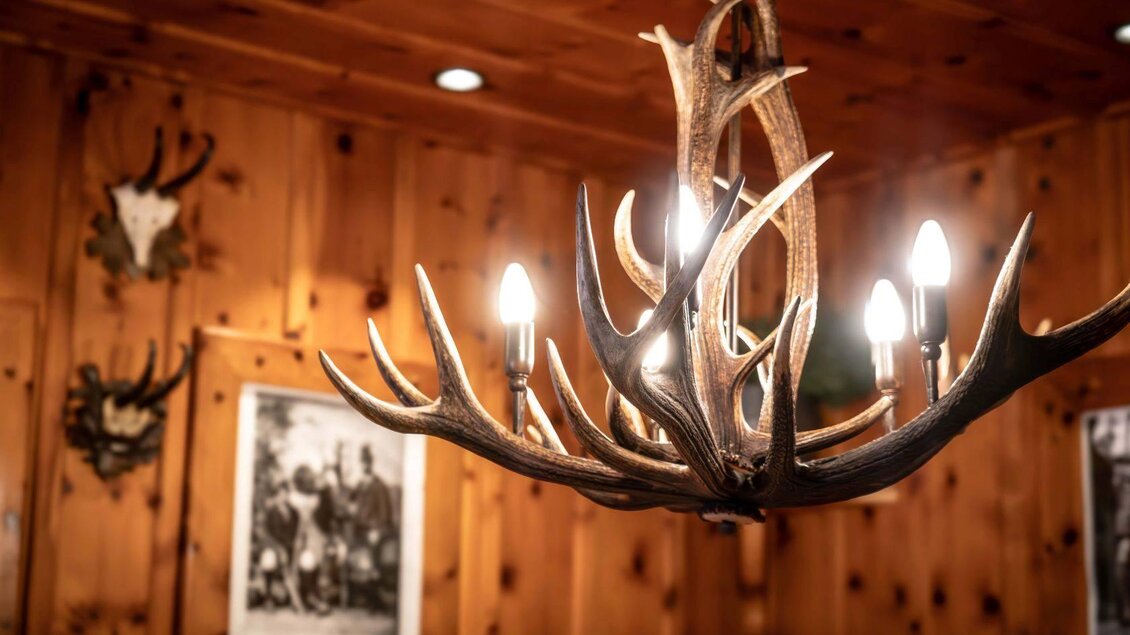 Eine rustikale Lampe aus Geweih mit mehreren Lichtquellen hängt in einem holzverkleideten Raum. Im Hintergrund sind Tierkopf-Dekorationen und alte Fotos an der Wand zu sehen. | © Chris Gütl