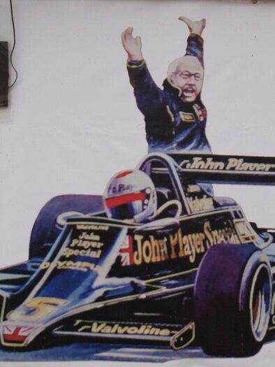 Eine bunte Wandmalerei eines Formel-1-Autos mit einem jubelnden Fahrer. Über dem Bild hängen zwei leuchtende „OPEN“-Schilder. | © Lotus Museum