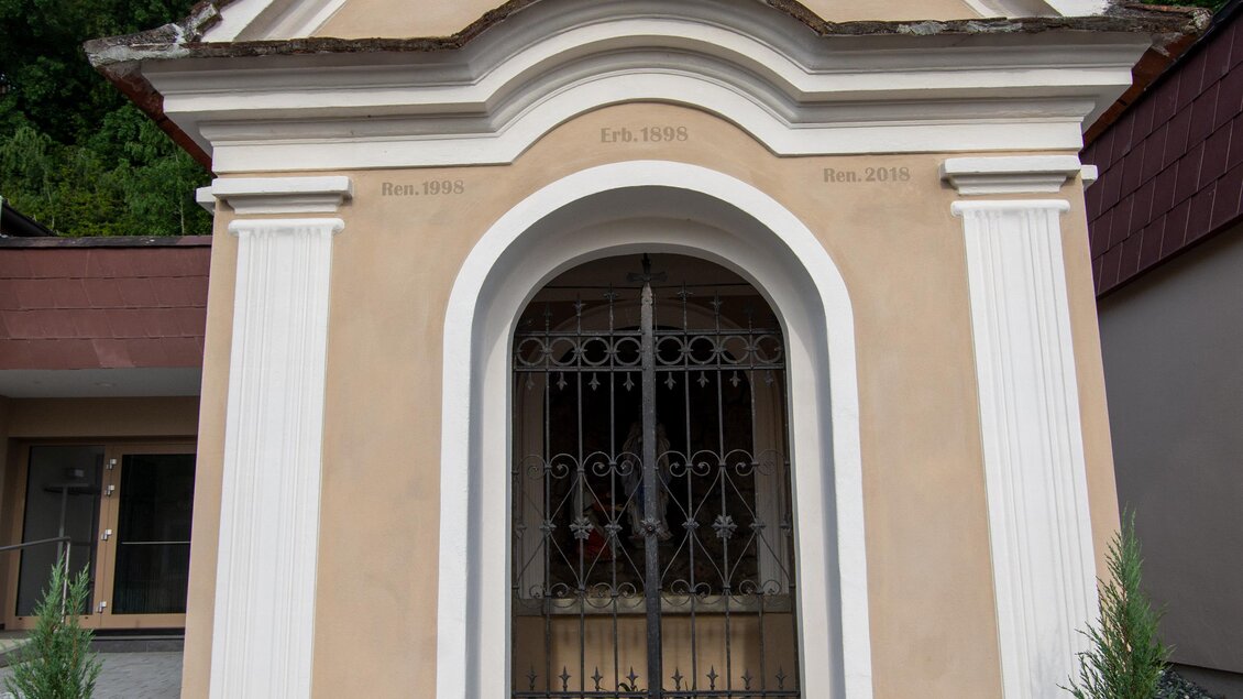 Eine kleine Kapelle mit einem zentralen Eingang und einer Kreuze. Es gibt grüne Pflanzen und einen Kiesweg davor. | © Kurkommission Bad Blumau