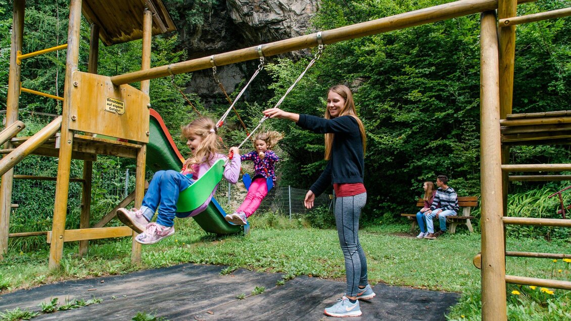 Ein Spielplatz mit Schaukeln in der Natur. Eine Frau schwingt zwei Kinder, während andere im Hintergrund sitzen. | © TV Region Graz - Mias Photoart