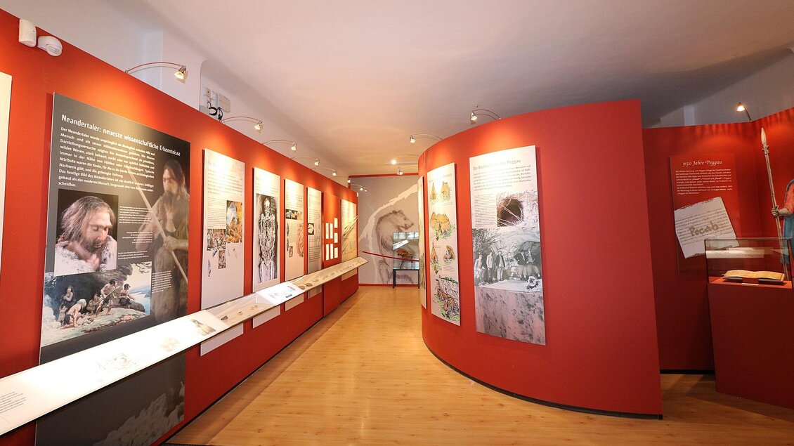 Ein Museum mit einer roten Wand, die mit informativen Tafeln und Bildern ausgestattet ist. Der Raum ist hell und einladend gestaltet. | © TV Region Graz - J. Lunghammer