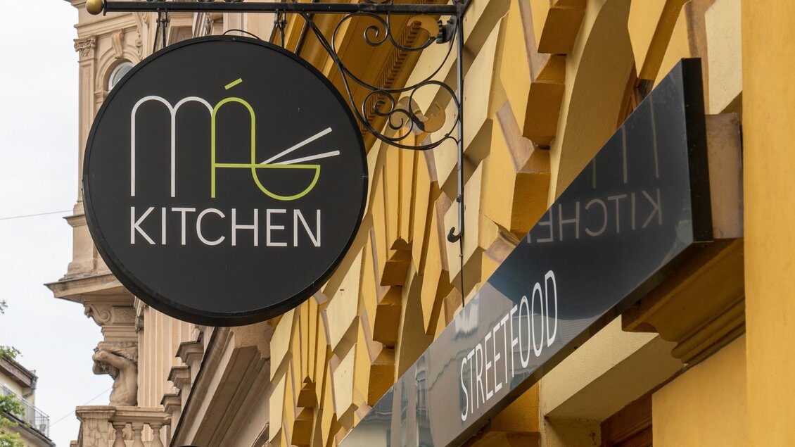 Schwarzes Rundschild mit Logo „Má Kitchen“ und Schriftzug „Streetfood“ an gelber Altbaufassade. | © Má Kitchen