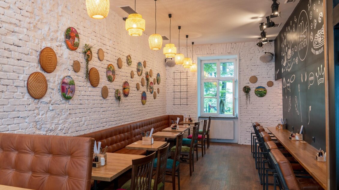 Langer Raum mit Lederbänken, bunten Stühlen, Wandkörben und Tafel mit Illustrationen. | © Má Kitchen
