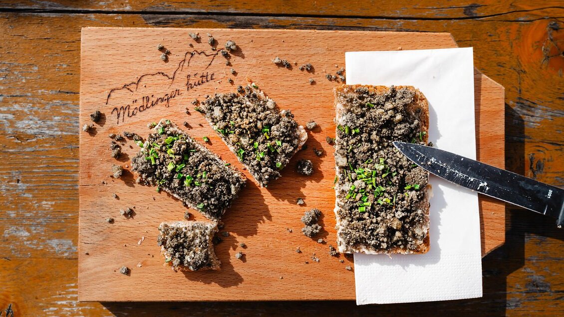 Ein Holzbrett mit einer Scheibe Brot, die mit einer dicken Schicht von dunkelgrauem Aufstrich und frischen Kräutern belegt ist. Neben dem Brot liegt ein Messer und ein Papiertuch.
