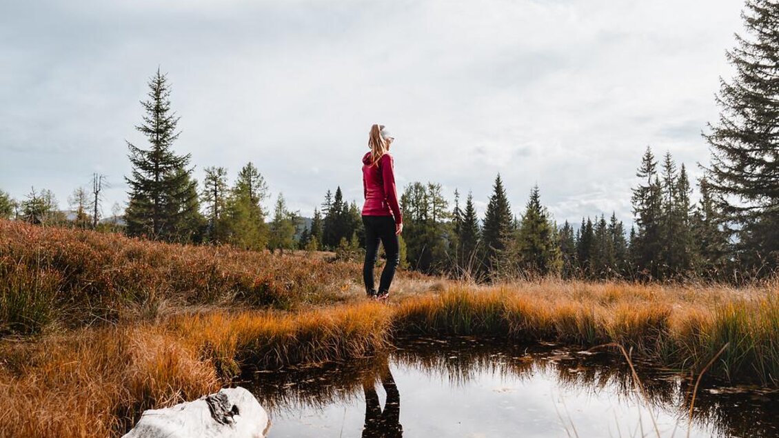 Eine Frau steht am Ufer eines kleinen Teichs in einer herbstlichen Landschaft. Die Umgebung ist von goldenen Gräsern und grünen Bäumen geprägt, unter einem bewölkten Himmel.