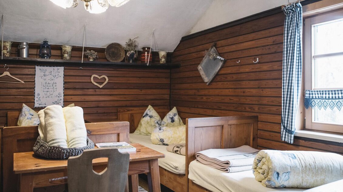 Ein gemütlicher Raum mit Holzverkleidung, zwei Betten und einem Esstisch. An der Wand hängen Erinnerungsstücke und eine Lampe sorgt für warme Beleuchtung. | © Stefan Leitner