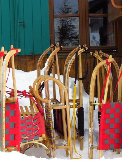 A group of wooden sleds stands in the snow, next to a wooden building. The sleds have red seats with a checkered pattern. | © Mörsbachwirt