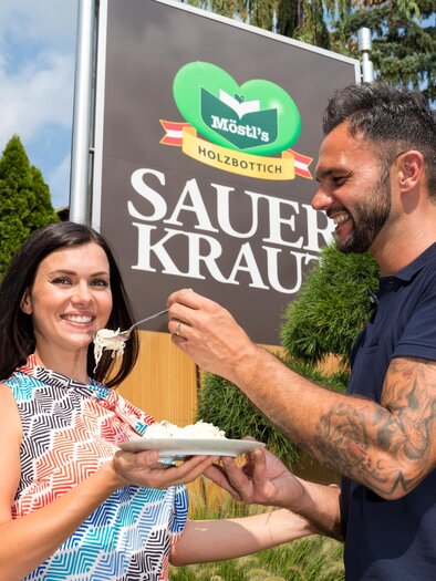 Ein Mann füttert eine Frau mit einem Löffel Sauerkraut vor einem Schild. Im Hintergrund sind grüne Bäume zu sehen. | © Region Graz - Harry Schiffer
