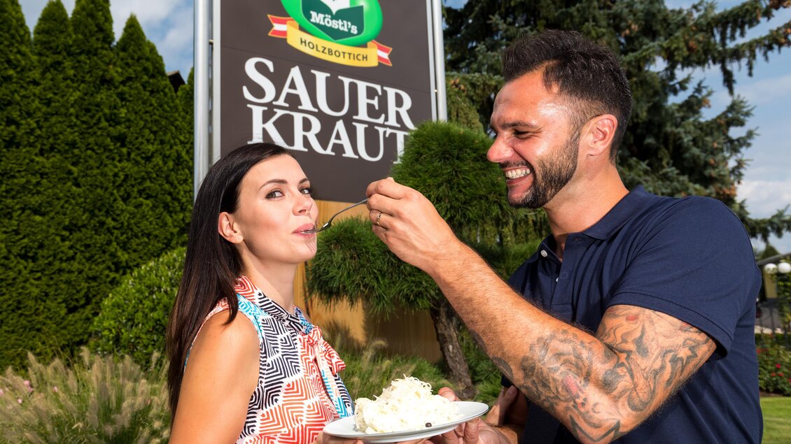 Ein Mann füttert eine Frau mit Sauerkraut vor einem Werbeschild. Im Hintergrund sind schöne Bäume zu sehen. | © Region Graz - Harry Schiffer