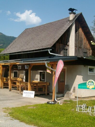 Ein gemütliches Gebäude mit Holzverkleidung und einem großen Dach. Draußen stehen Tische mit Sonnenschirmen in einer grünen Landschaft.