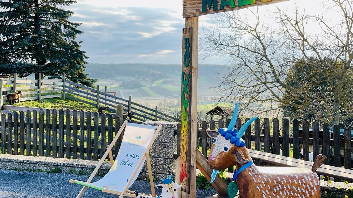 Ein malerischer Aussichtspunkt mit einer bunten Holzstruktur namens „MALDUHOF“. Im Vordergrund steht ein hölzernes Ziegenfigur und ein Liegestuhl.