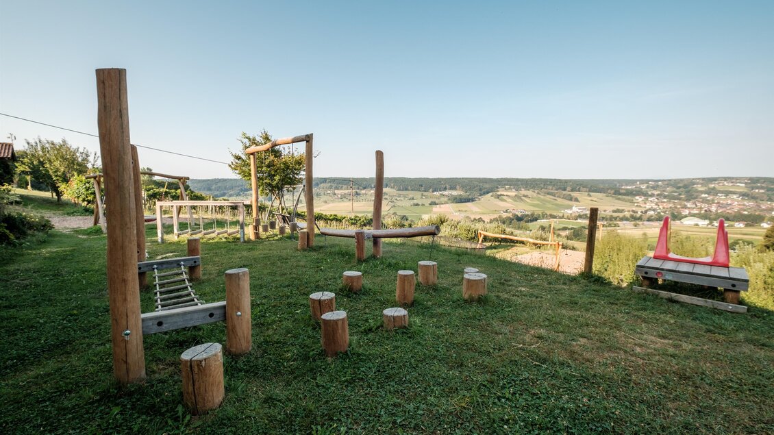Ein Spielplatz mit Holzgeräten und einer Aussicht auf die Landschaft. Der Himmel ist klar und die Umgebung sieht grün und einladend aus. | © MALU