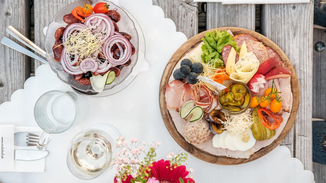 Ein geschmackvoll angerichteter Tisch mit zwei Platten: einer mit Salat und Zwiebeln sowie einer mit verschiedenem Fleisch, Käse und Gemüse. Dazu ein Glas Wasser und eine Blumenvase. | © MALU