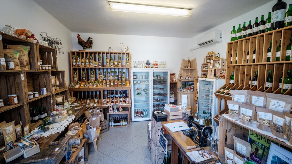 Ein gemütlicher Laden mit Regalen aus Holz, die verschiedene Produkte und Lebensmittel anbieten. An der Wand stehen Flaschen und Gläser, und es gibt eine Kühltruhe für frische Waren. | © MALU