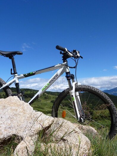 A mountain bike is standing on a large stone in a picturesque landscape. The sky is clear and blue, with some clouds in the distance. | © Pixabay