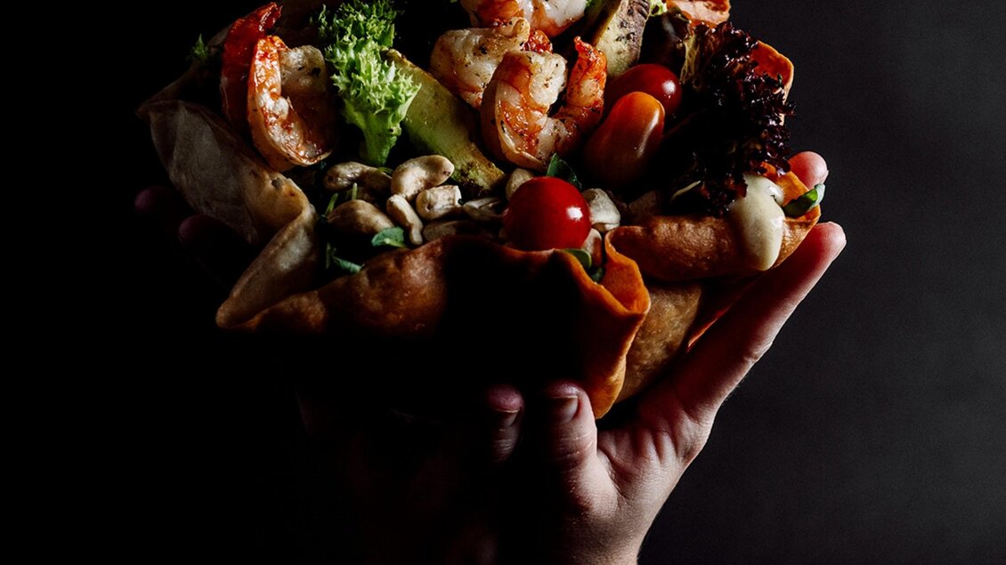 Hand hält eine mit Garnelen, Salat, Avocado und Nüssen gefüllte Tortilla-Bowl. | © MEAT.art - Simona Donko