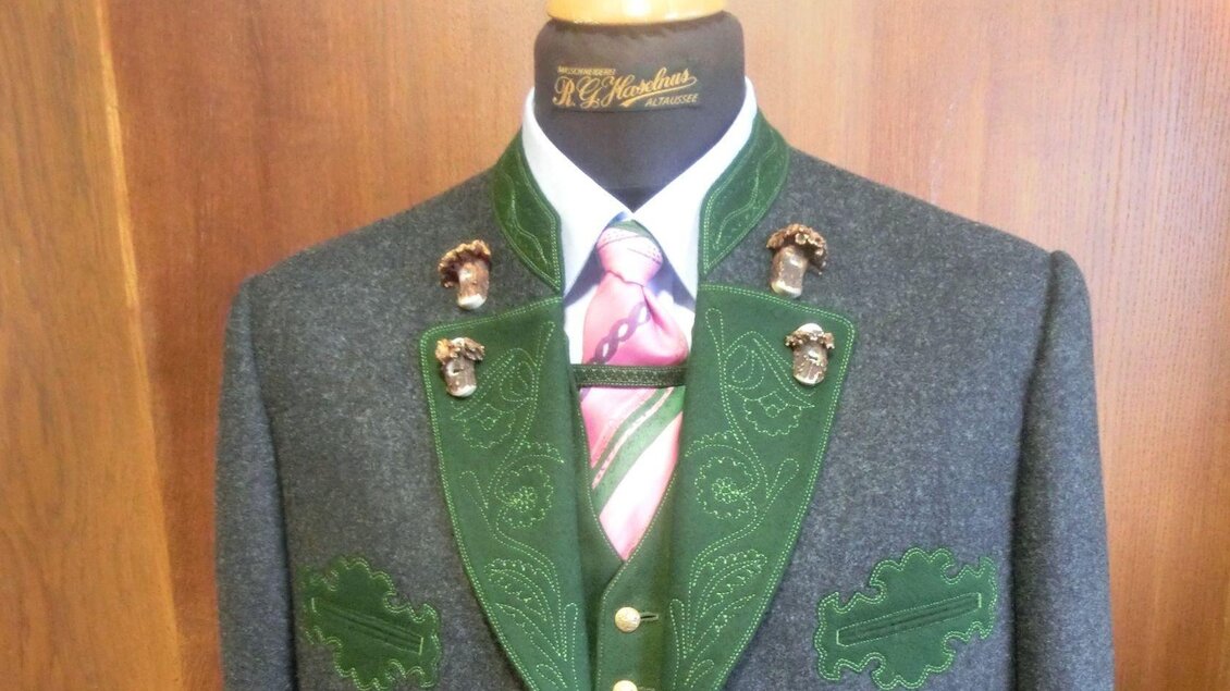Ein traditioneller Trachtenanzug mit einem grauen Stoff und grünen Applikationen. Das Outfit ist mit einer eleganten Krawatte und einem Hemd kombiniert. | © Haselnus Rene